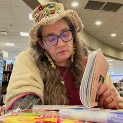 Jolynn reading a book at a bookstore, wearing a crocheted flower hat.