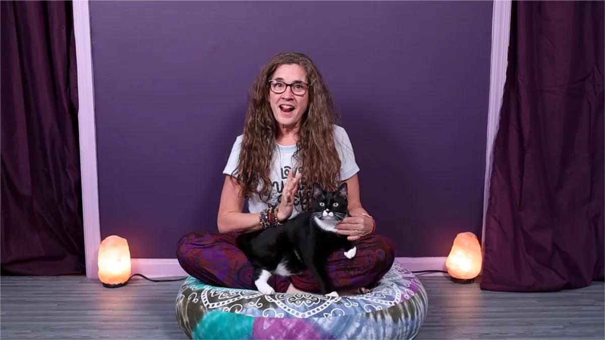 Jolynn sitting cross legged on a meditation cushion, laughing, with Miss Muffin the cat sitting on her lap.