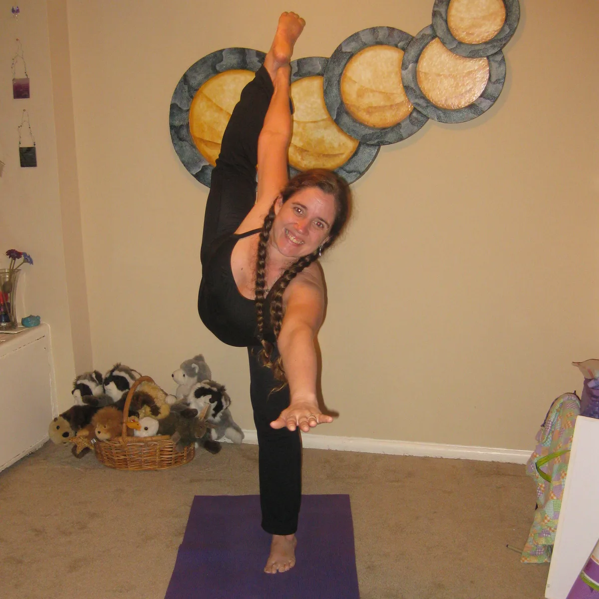 Jolynn doing a standing yoga pose, one leg raised straight up, in front of an abstract painting by Thad.
