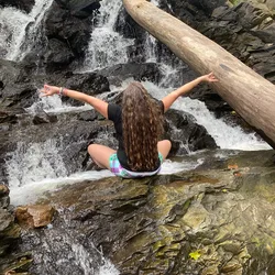 Jolynn sitting cross legged on rocks at a waterfall near Harpers Ferry, arms raised in a meditation mudra.